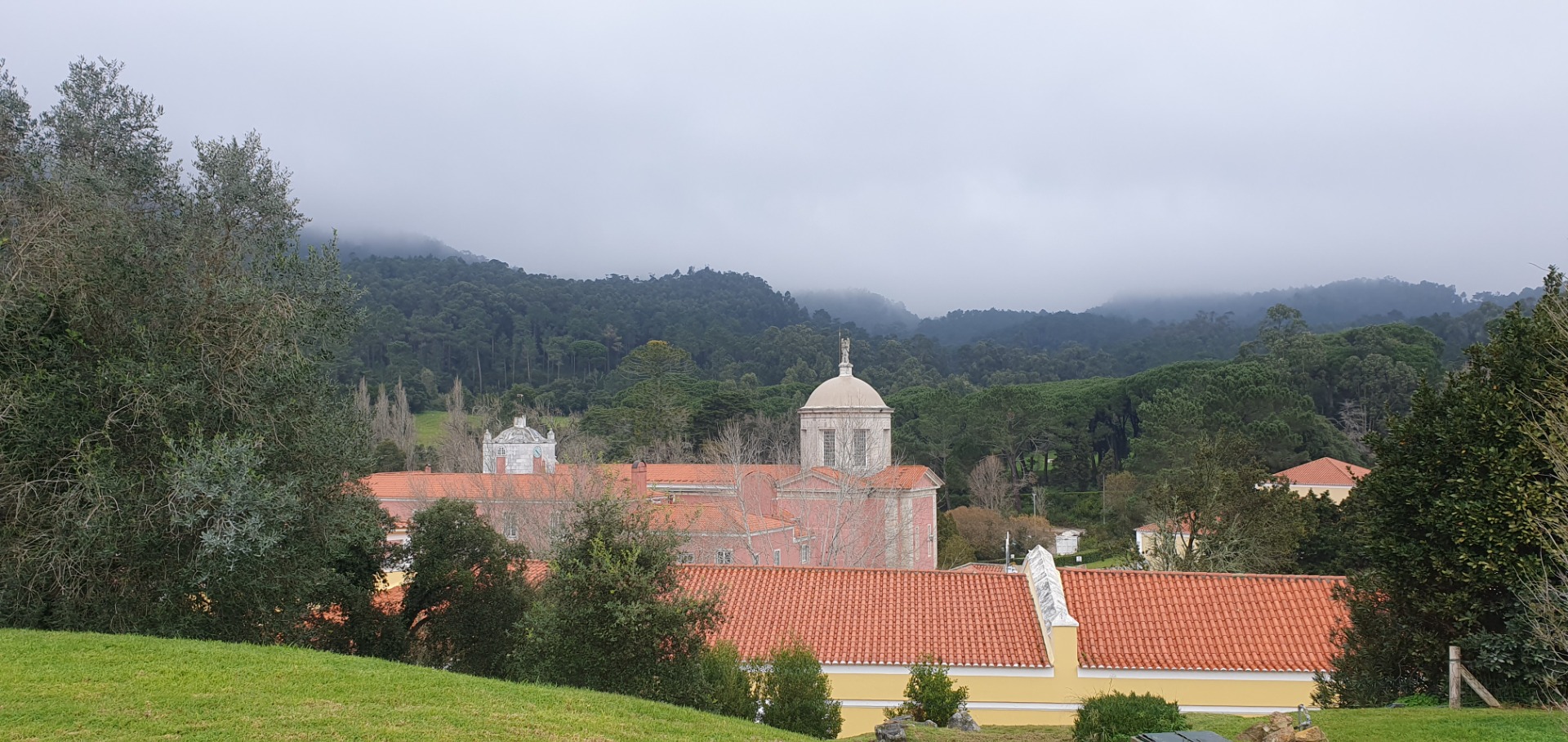 Historisches Klostergebäude mit Kuppel in Penha Longa vor bewaldeten Hügeln unter nebligem, wolkenverhangenem Himmel.