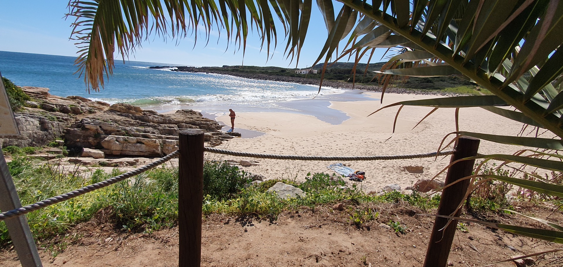 Blick auf einen einsamen Strand an detr Algarve mit Wellen, Felsen und einer Person unter Palmen, Meer im Hintergrund.