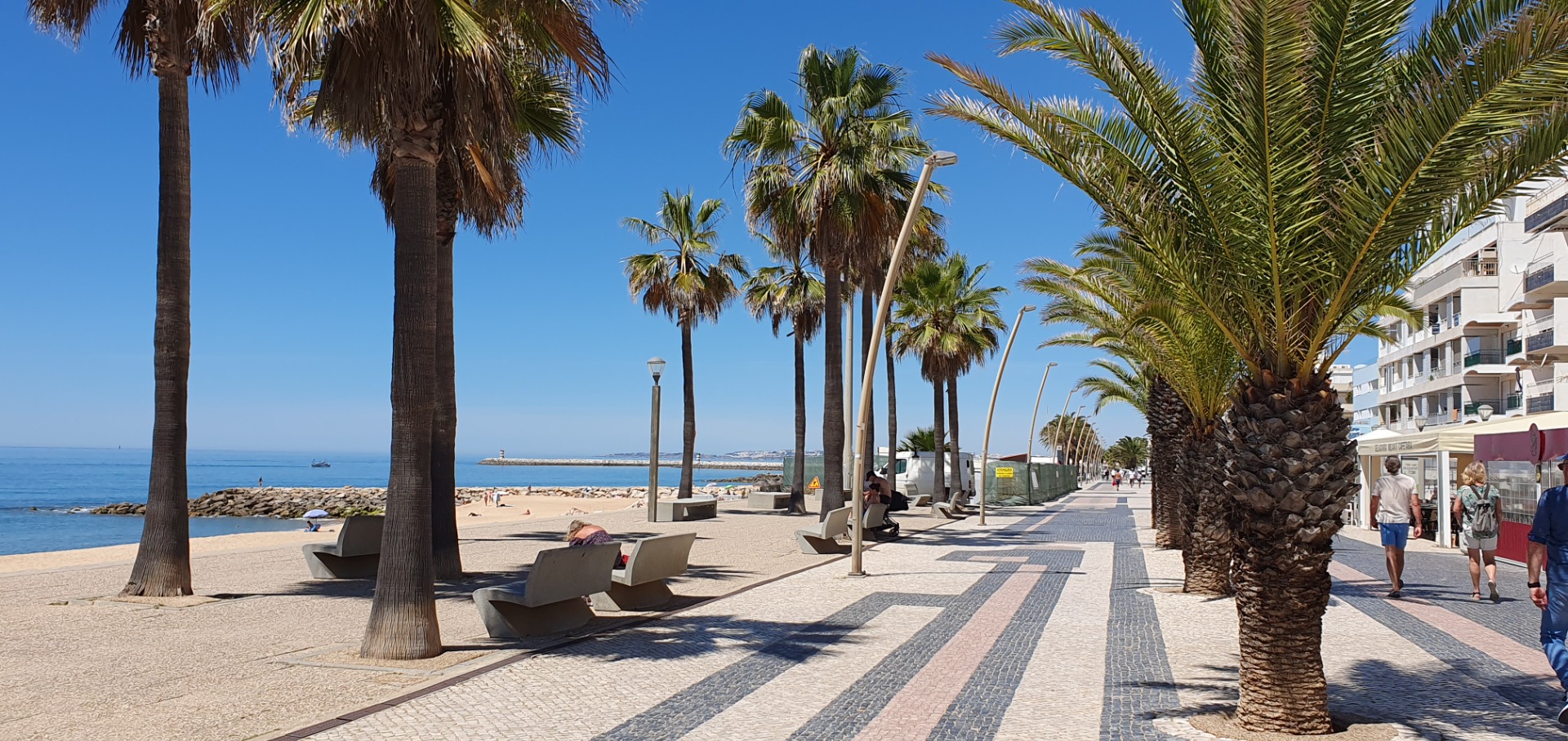 Sonnige Promenade Promenade am Strand von Quarteira - Algarve am Meer mit Palmen, Bänken, Laternen und vorbeigehenden Touristen vor Hotelgebäuden.
