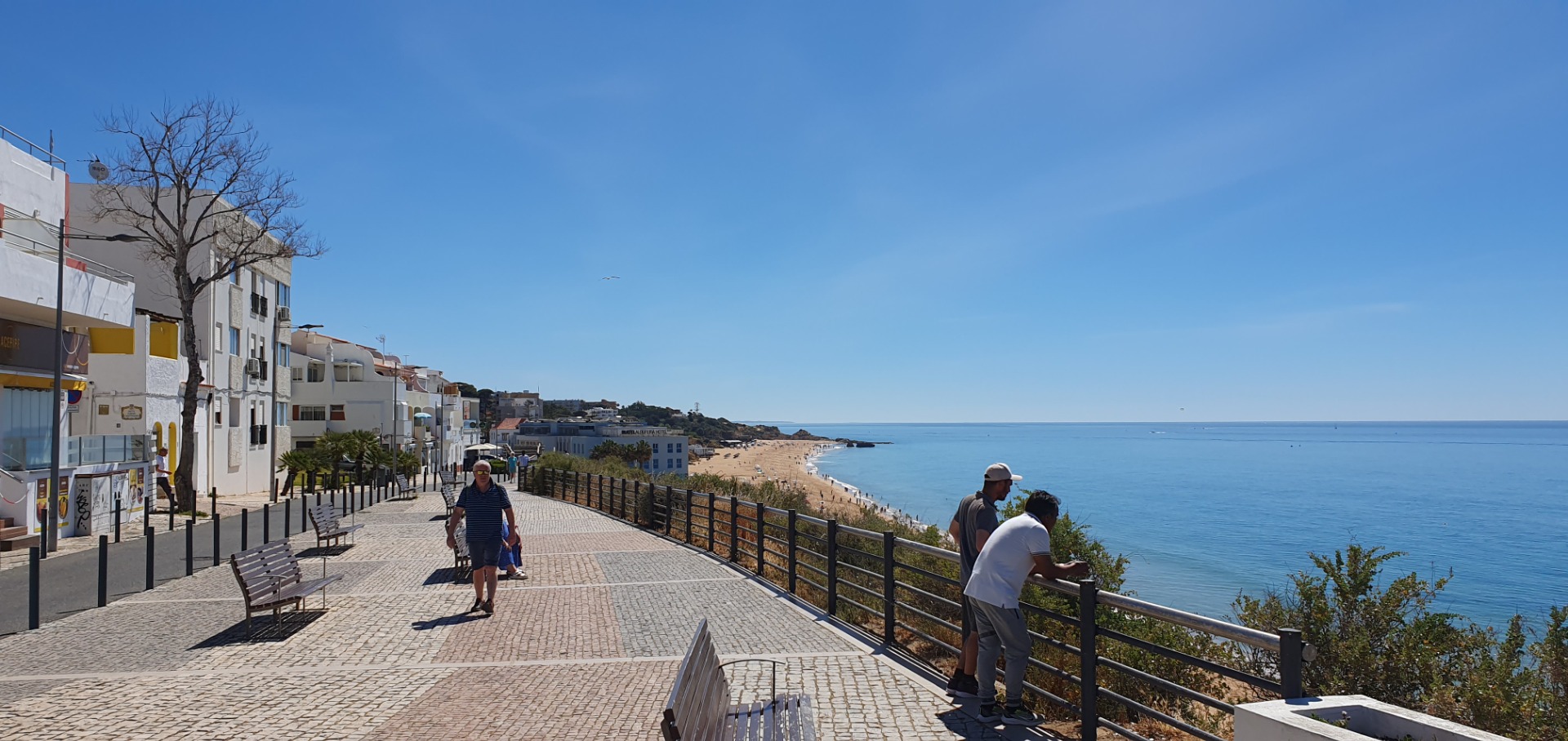 Promenade am Meer mit Spaziergängern, gepflastertem Weg, Bänken und Häusern entlang der Küste unter klarem Himmel.