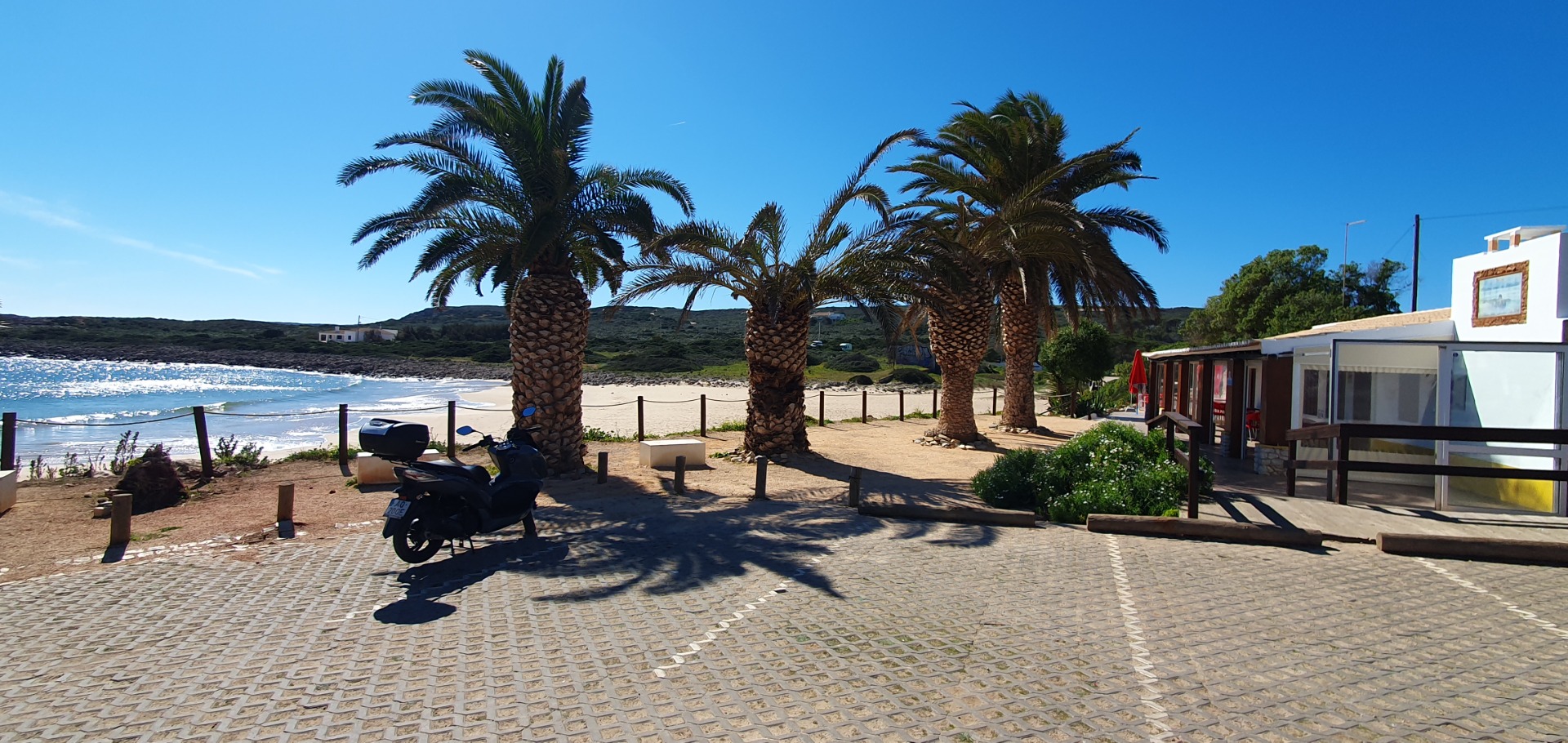 Promenade bei Praia da Luz Algarve mit Palmen, gepflastertem Weg, Motorrädern und weißen Gebäuden am Meer unter klarem blauen Himmel.