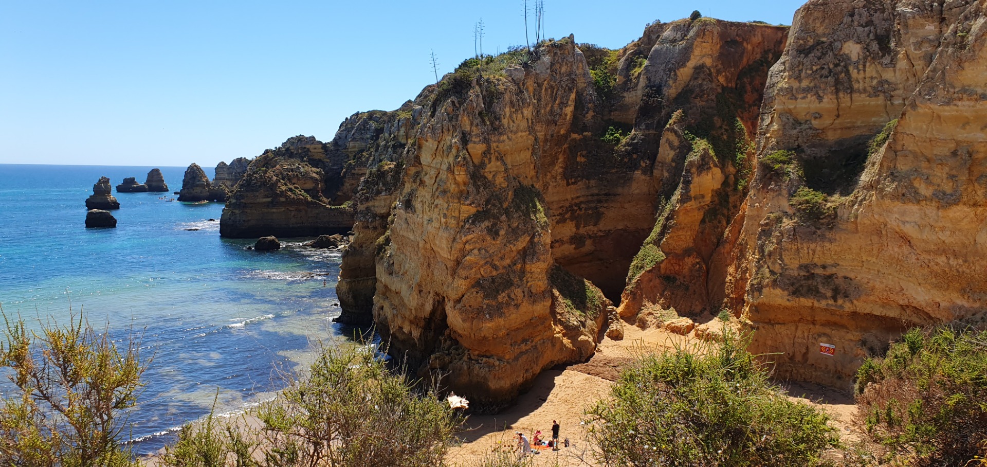 Steile orangefarbene Klippen an der Küste, türkisblaues Meer, kleine Felsinseln und Wanderer am Strand.