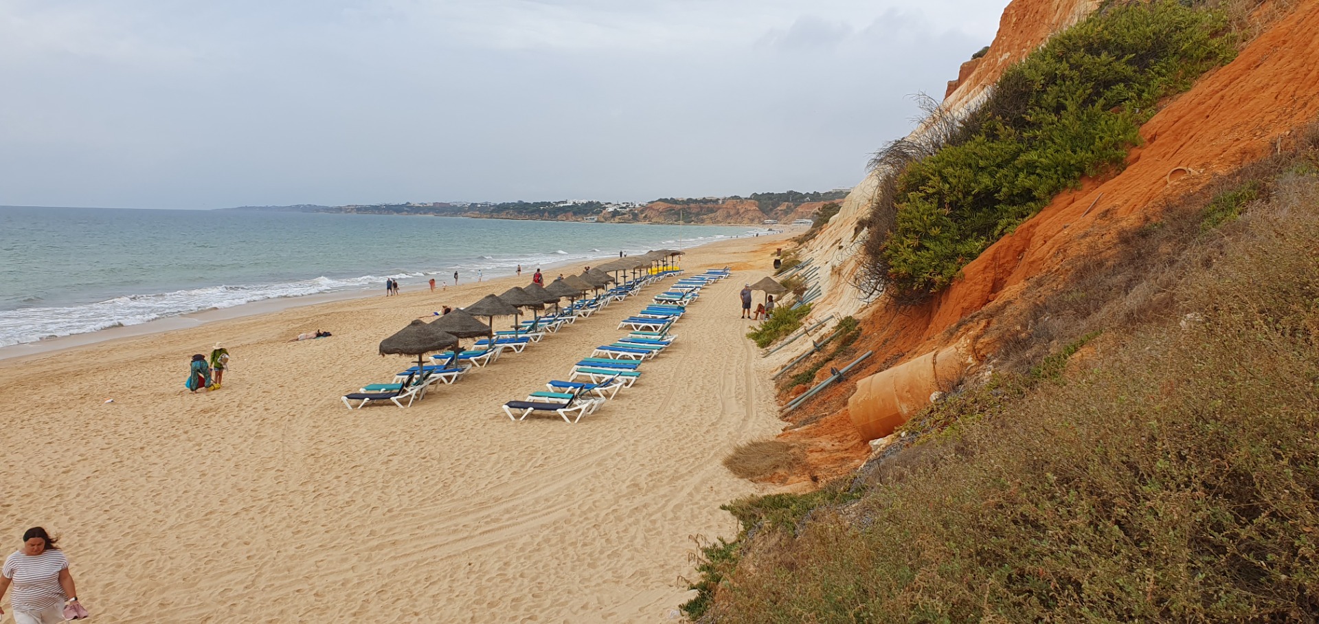 Sandiger Strand Praia da Falesia - Zentral Algarve mit blauen Liegen, Sonnenschirmen und türkisfarbenem Meer unter bewölktem Himmel.