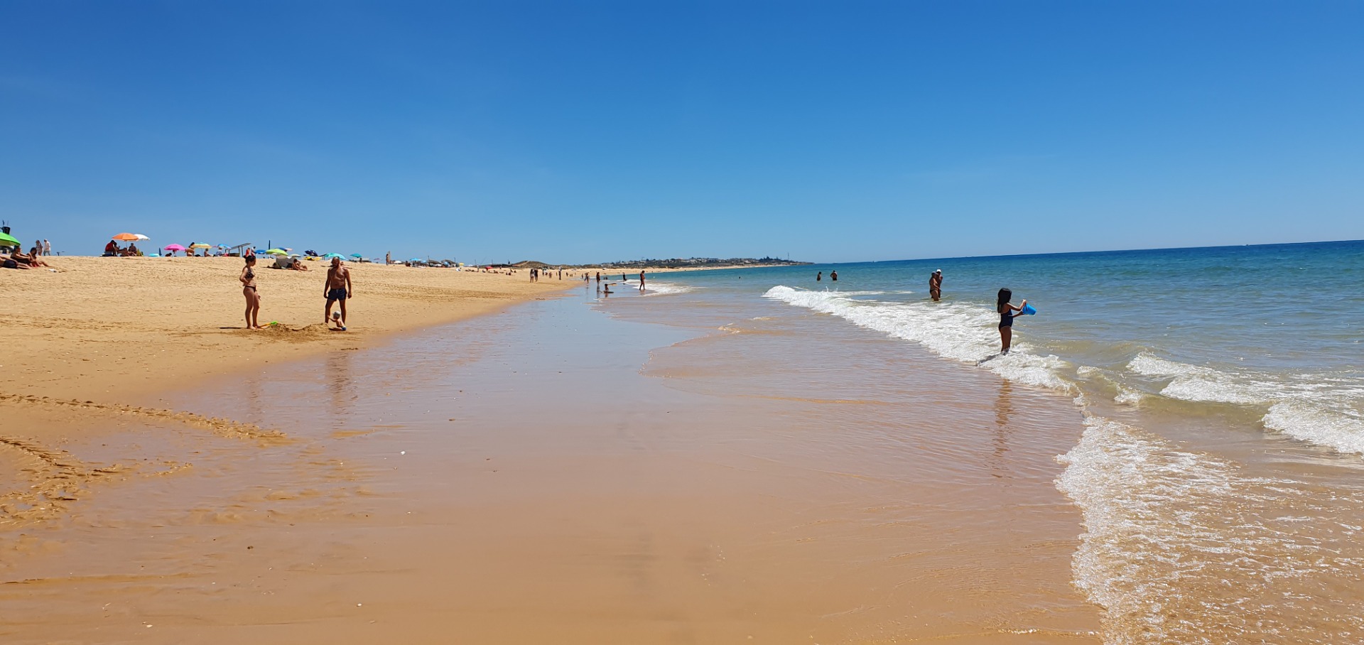 Breiter Sandstrand an der Algarve mit Badegästen, bunten Sonnenschirmen und ruhiger Brandung unter klarem blauen Himmel.