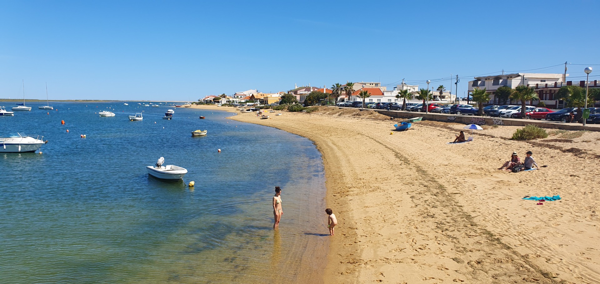 Sandstrand bei Faro Algarve mit flachem Wasser, mehreren Booten und Menschen beim Schwimmen, Häusern und Palmen am Ufer.