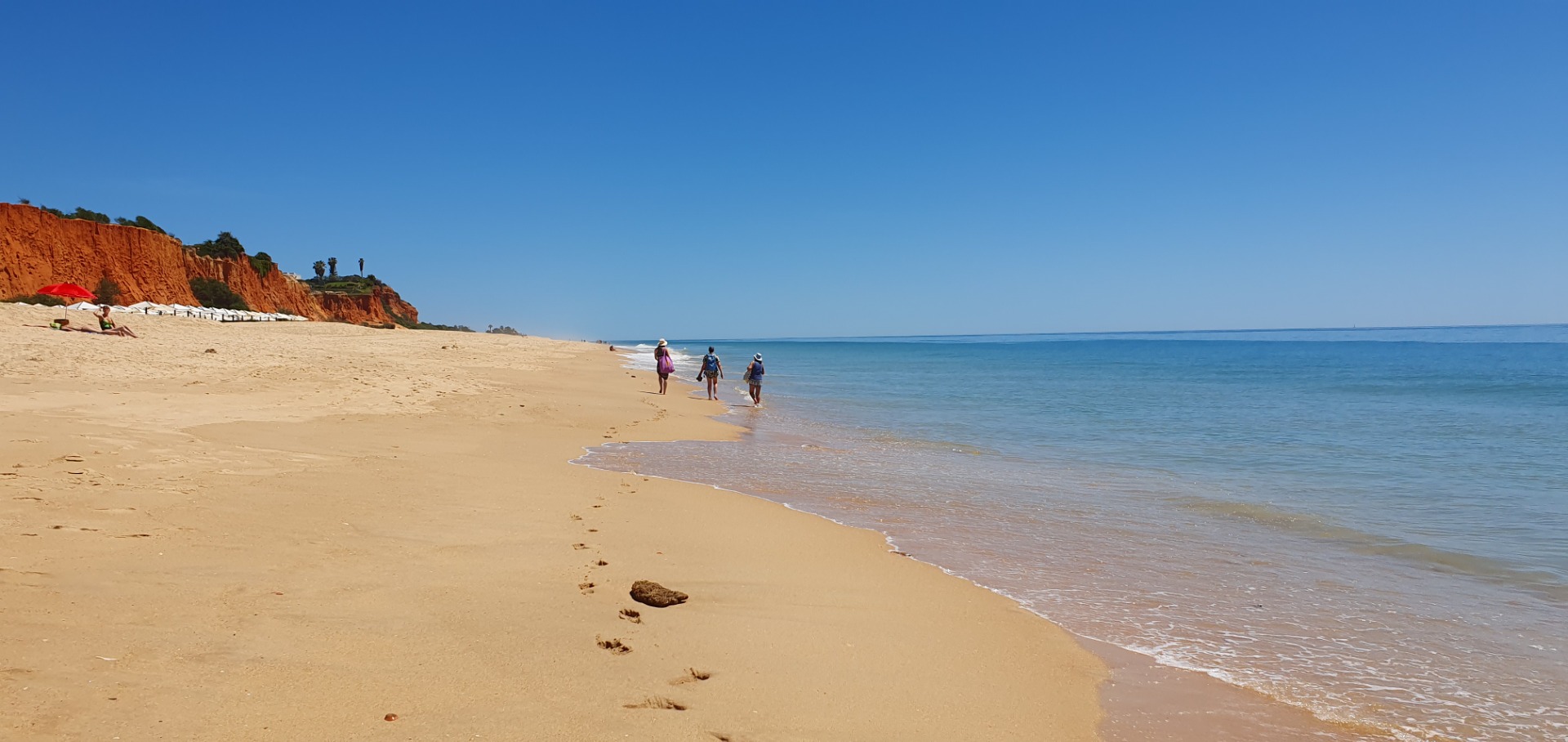 Sandiger Strand mit Spaziergängern, roter Klippe im Hintergrund und ruhigem blauen Meer unter klarem Himmel.