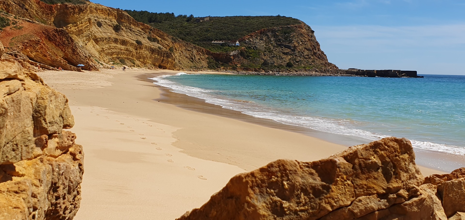 algarve Sandstrand mit türkisfarbenem Wasser, Klippen und grünen Hügeln, im Hintergrund ein kleiner Leuchtturm.