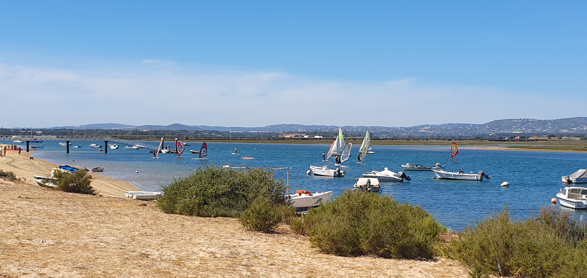 Segelboote und Windsurfer auf ruhigem See, Boote am Ufer, sonnige Landschaft mit Hügeln im Hintergrund.