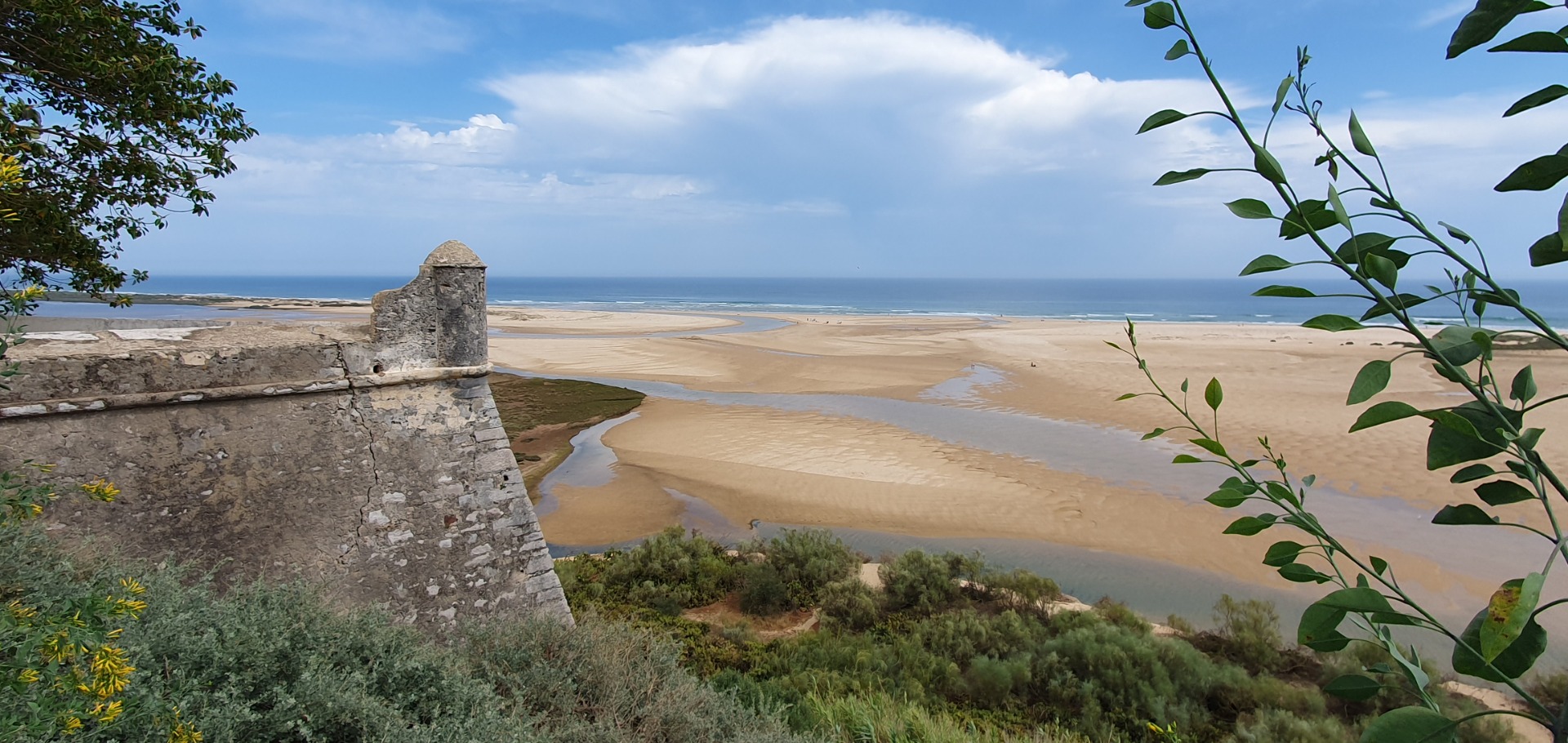 Felsruine am Strand mit Blick auf weite Sandbucht, Flussmündung und Meer unter bewölktem Himmel.