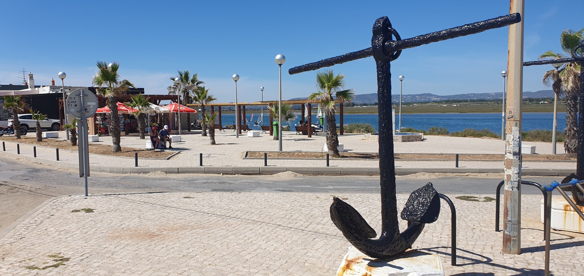 Schattige Promenade am Meer mit Palmen, Sonnenschirmen, Sitzgelegenheiten und einer großen Anker-Skulptur im Vordergrund.