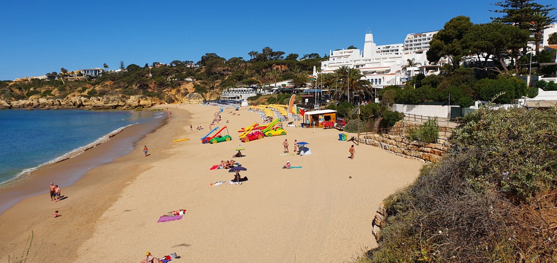 Sandstrand mit bunten Liegen, Surfbrettern und Menschen, daneben weiße Hotelgebäude und Bäume unter blauem Himmel.