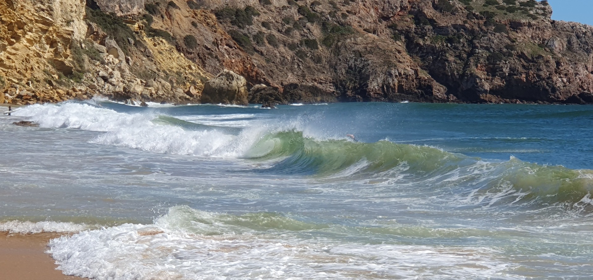 Grüner Surfer-Wave bricht an felsiger Küste, Schaum und Spritzer im Vordergrund, sonniger Tag.