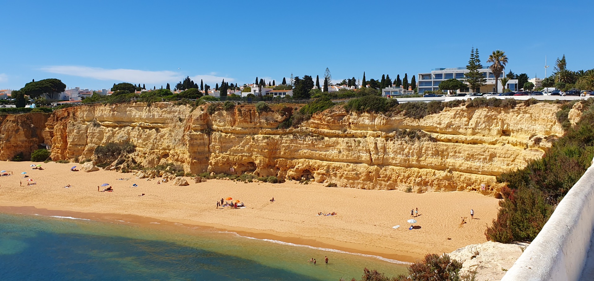 Sandstrand mit Badegästen, türkisfarbenem Meer, steilen Felsklippen und Küstenhäusern unter blauem Himmel.
