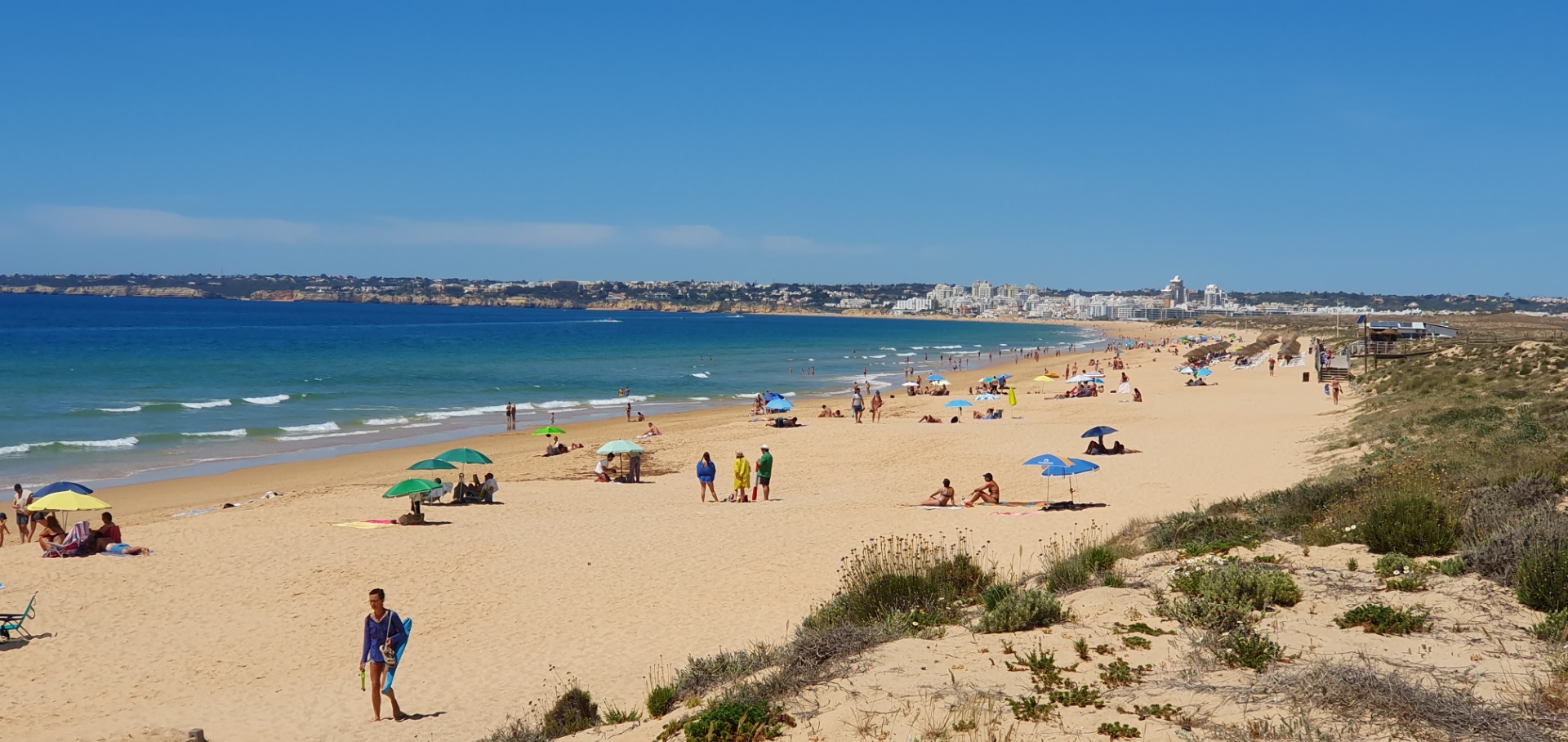 Breiter Sandstrand mit vielen Sonnenliegen, bunten Schirmen, badenden Menschen und Küstenstadt in der Ferne.
