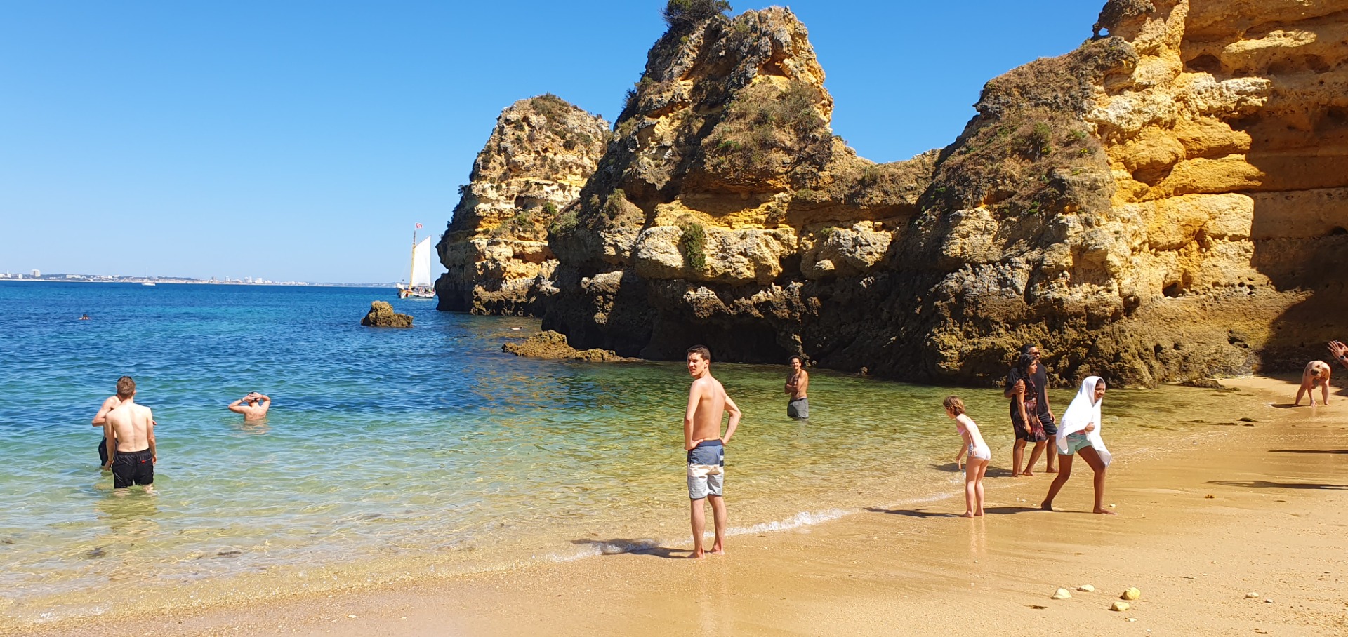 Menschen stehen am Strand im klaren Wasser vor hohen Felsklippen, Sonne und blauer Himmel.