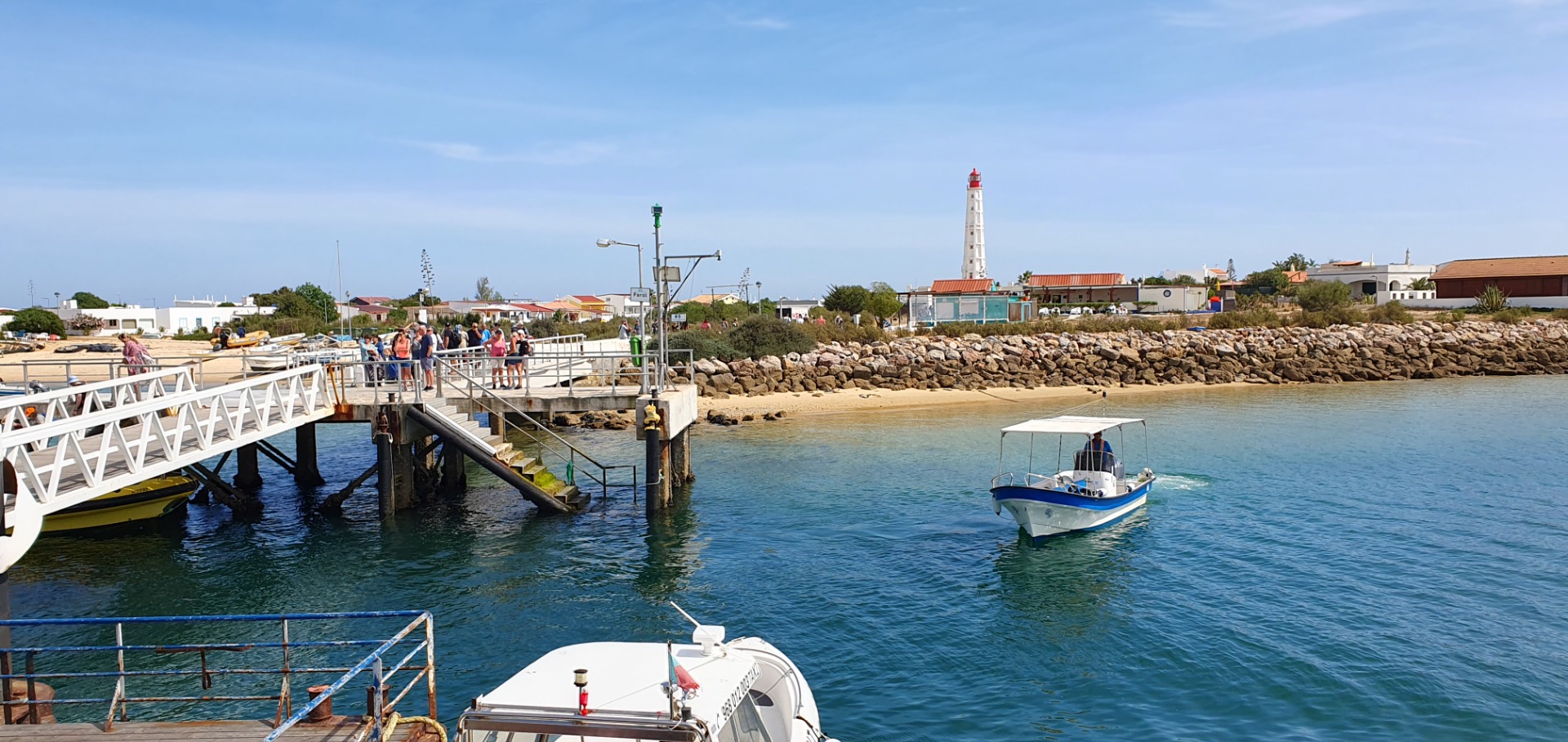 Hafenpromenade mit weißem Steg, Leuchtturm, Booten im Wasser und Spaziergängern bei klarem Himmel.