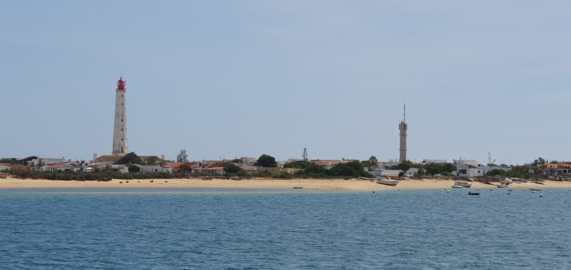 Weißer Leuchtturm mit roter Spitze und Hafenanlagen am Strand, Boote im Vordergrund, ruhiges Meer.