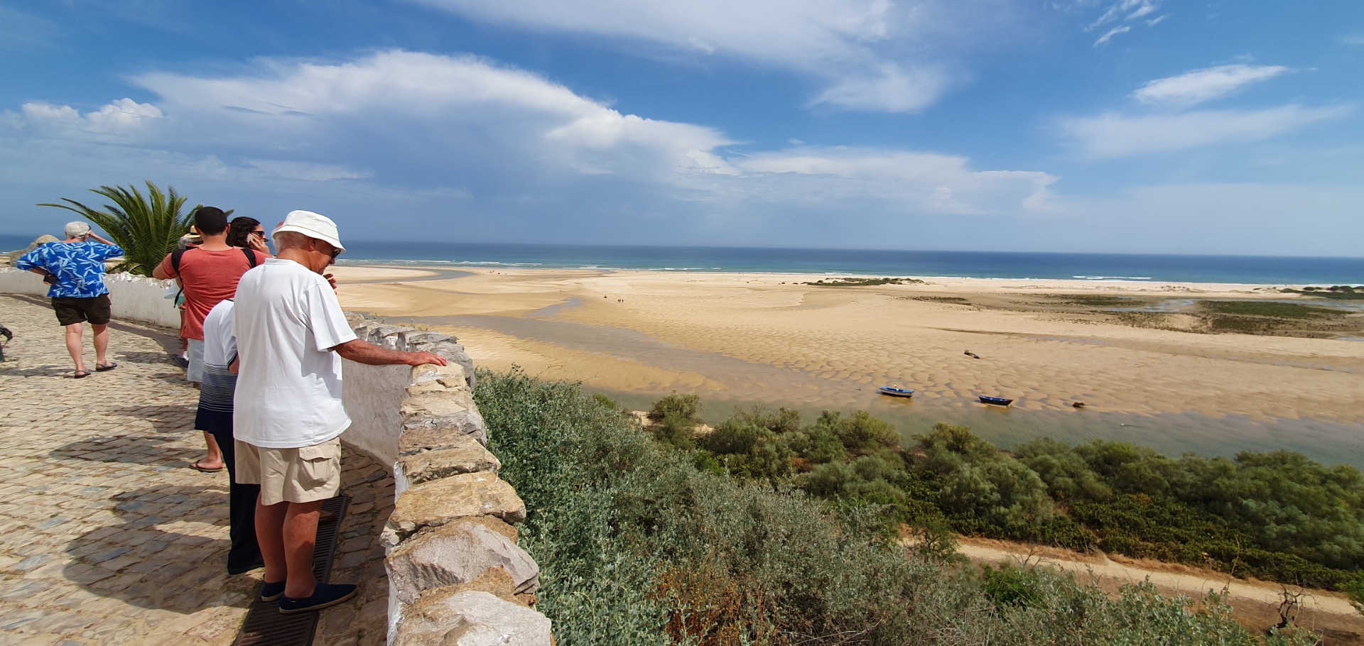 Touristen stehen an einer Klippe und blicken auf einen weiten Sandstrand mit Booten und blauem Meer.