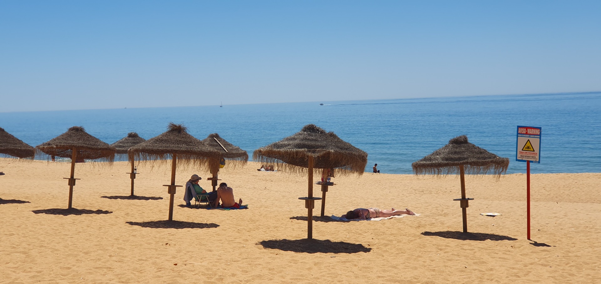 Sandiger Strand mit mehreren Strohdächern, Liegen und Badegästen vor ruhigem Meer und blauem Himmel.