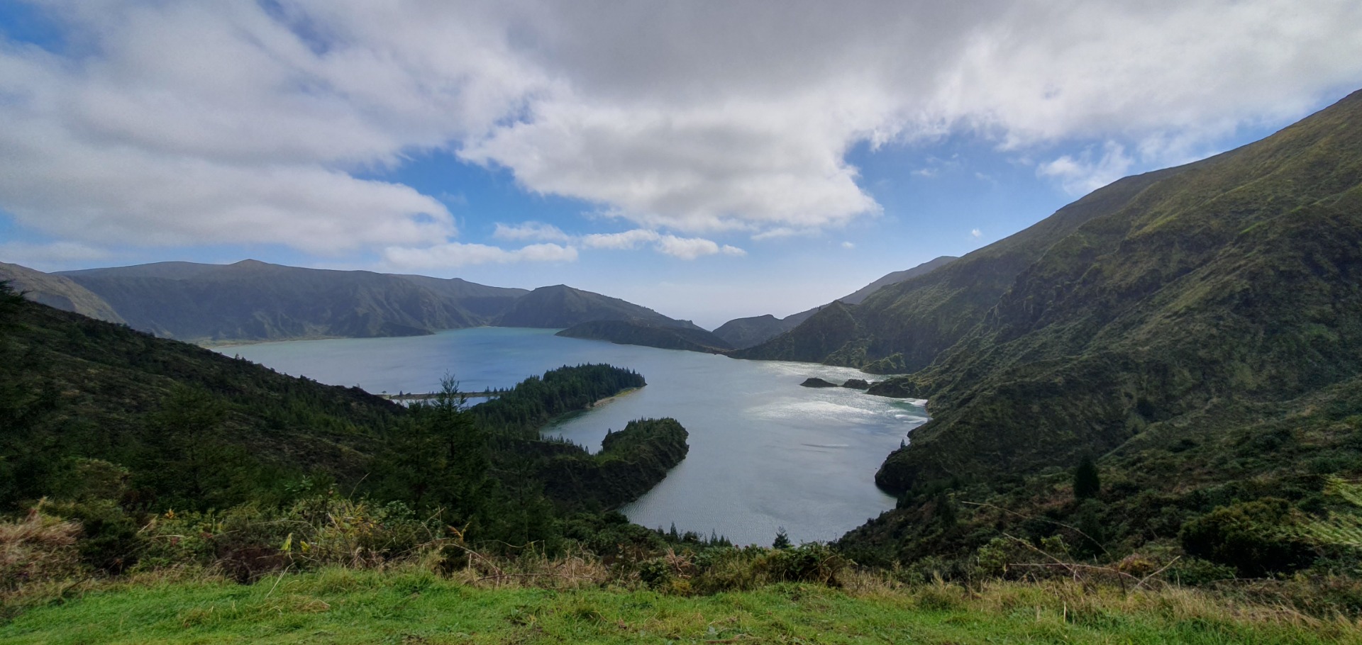 Breiter Fjord zwischen grünen Bergen, ruhiges türkisfarbenes Wasser, Wolken am Himmel, Blick von einem Hang.