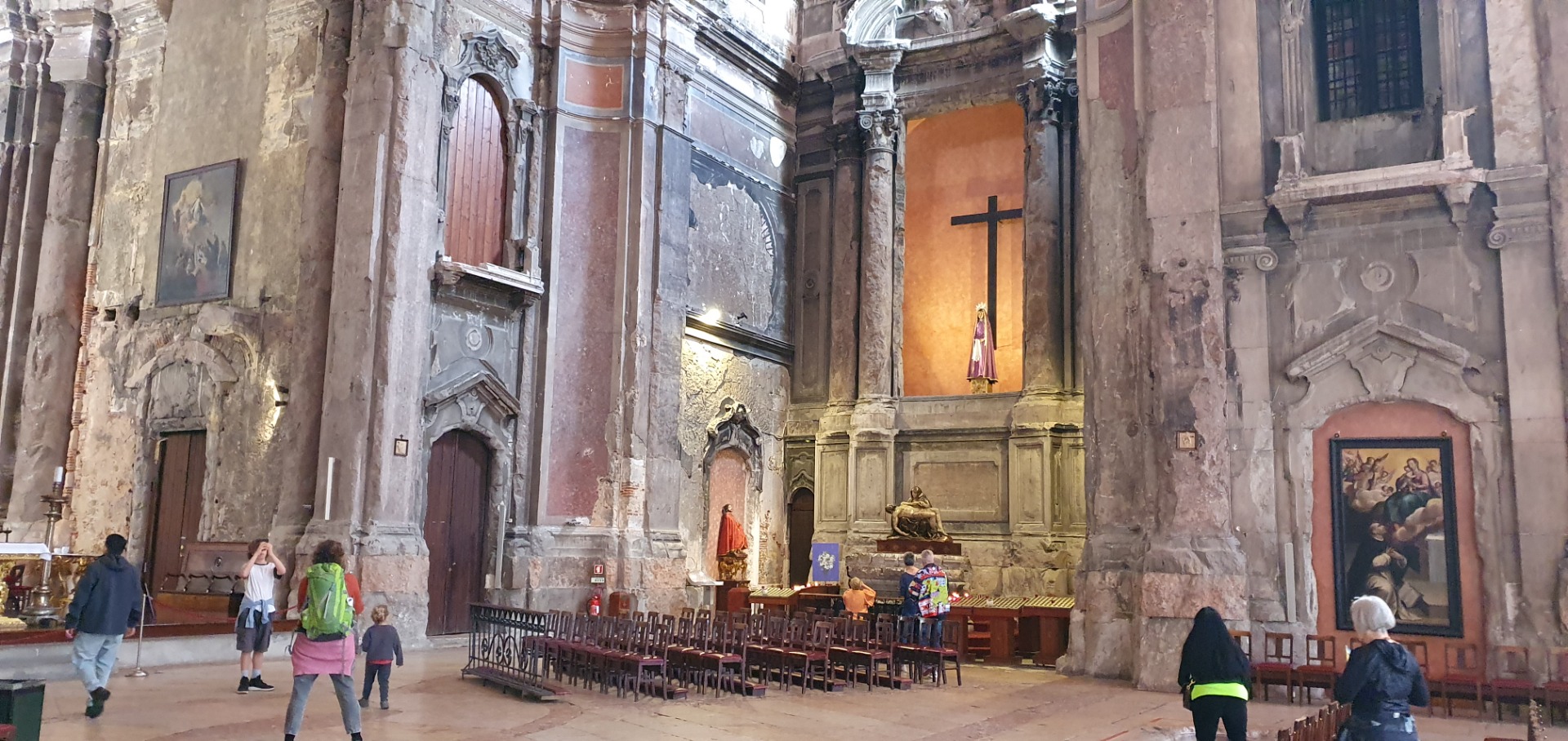 Innenraum einer historischen Kirche mit Altar, goldener Statue, Kreuz und Besuchern, umgeben von hohen Säulen.