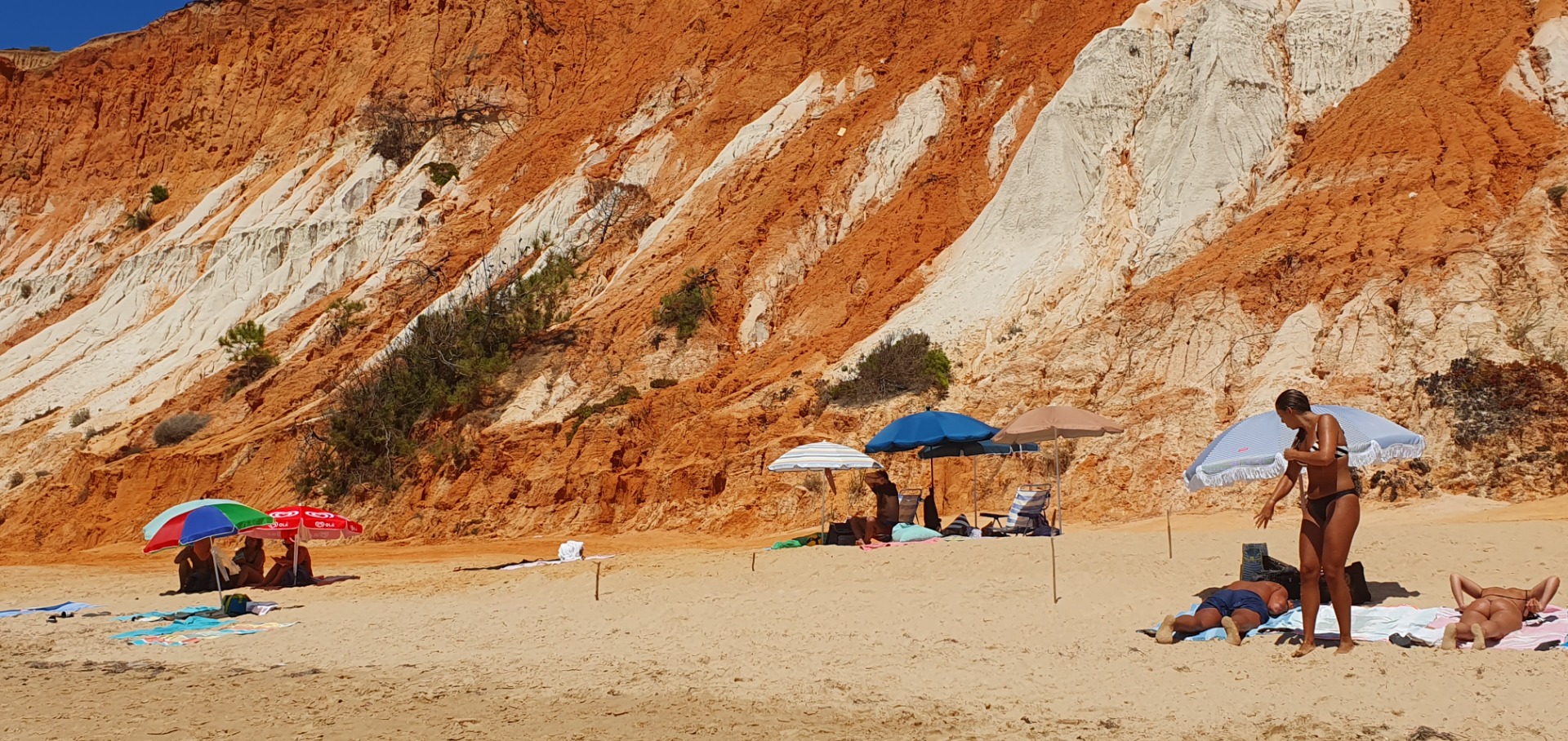 Beachgoers relax under umbrellas on sandy shore beneath towering red and white canyon cliffs.