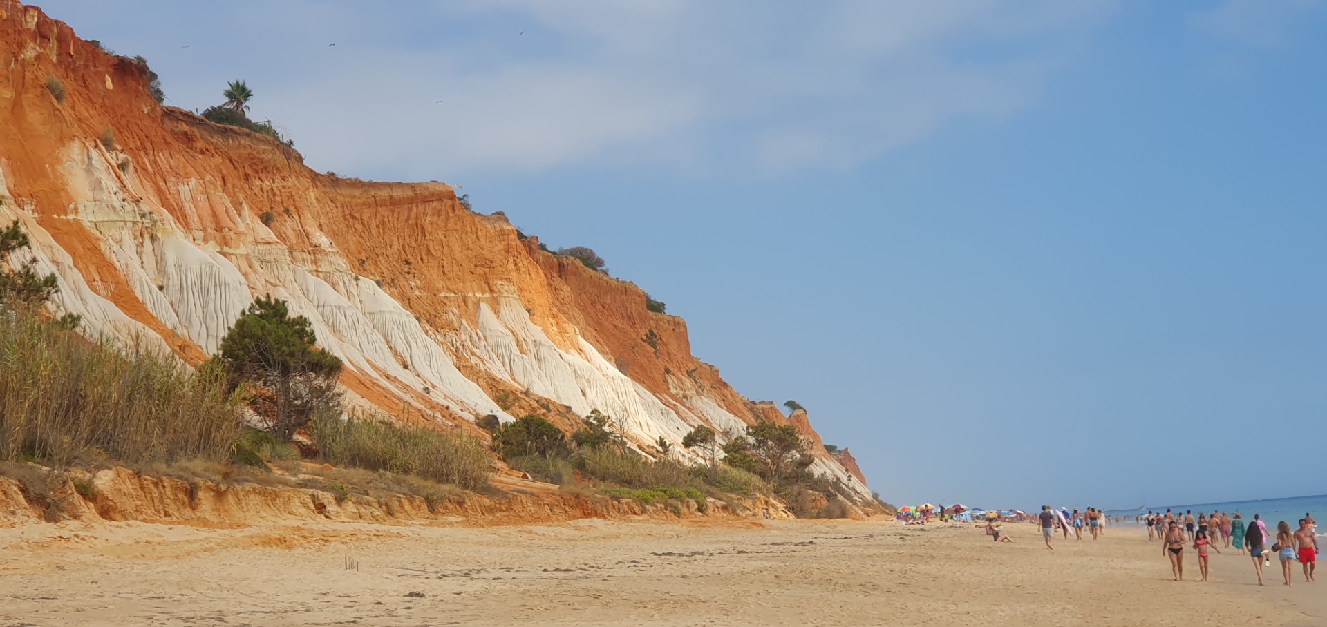 Sunny beach with people walking past orange-and-white coastal cliffs under a blue sky.