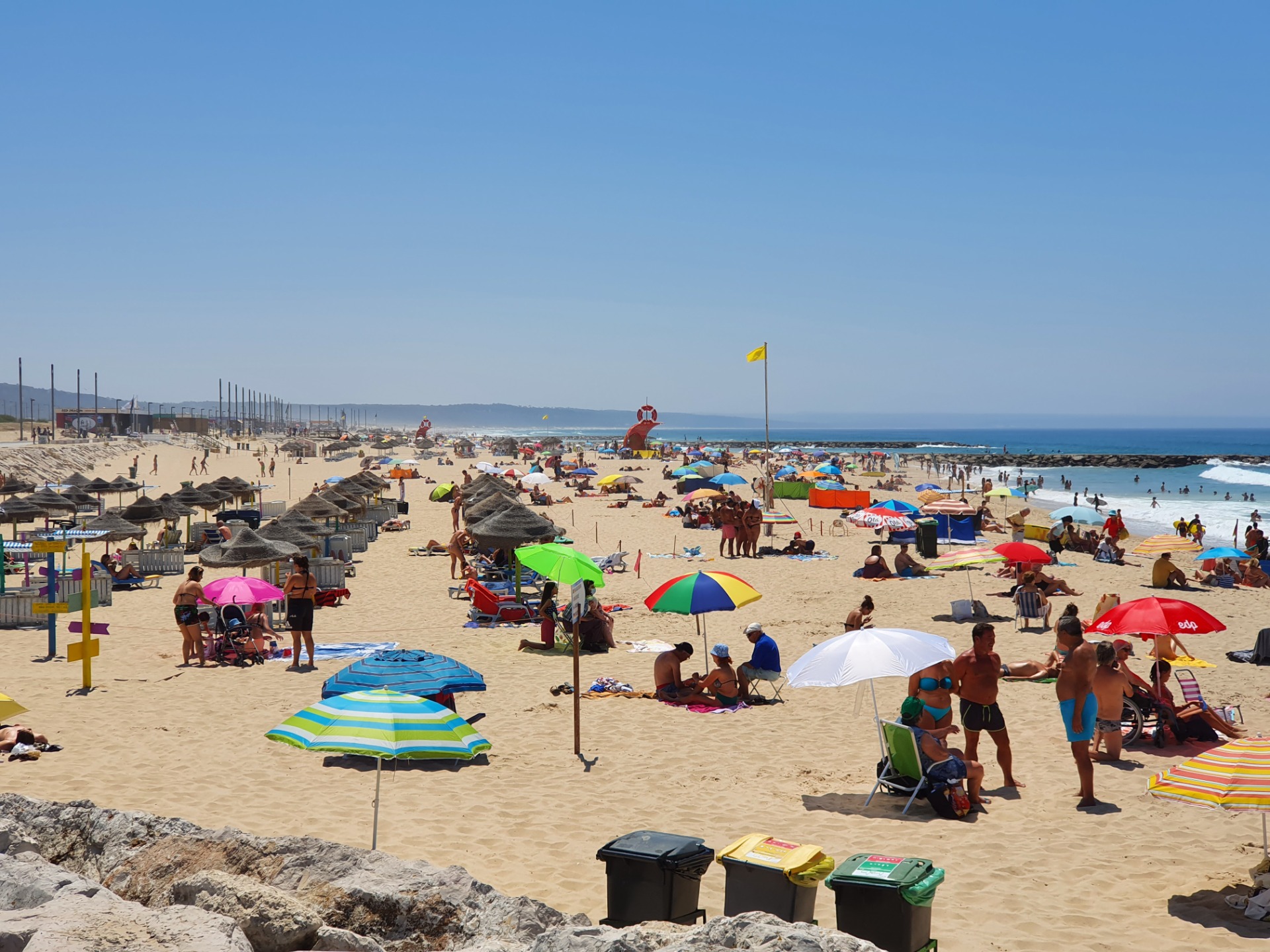 Crowded Costa da Caparica Beach sandy beach with colorful umbrellas, sunbathers, and ocean waves under a clear blue sky.