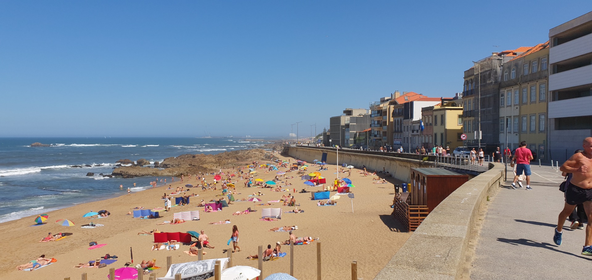 Porto, Sunny seaside beach with umbrellas and swimmers, promenade buildings along the right, clear blue sky.