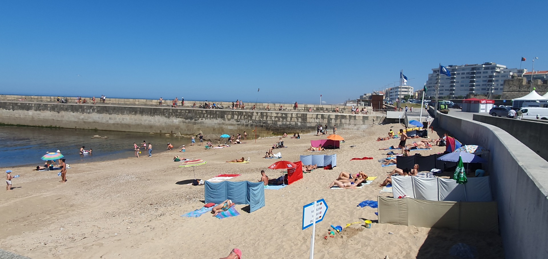 Sunny beach scene with people relaxing, tents and umbrellas along a seawall under clear blue sky.