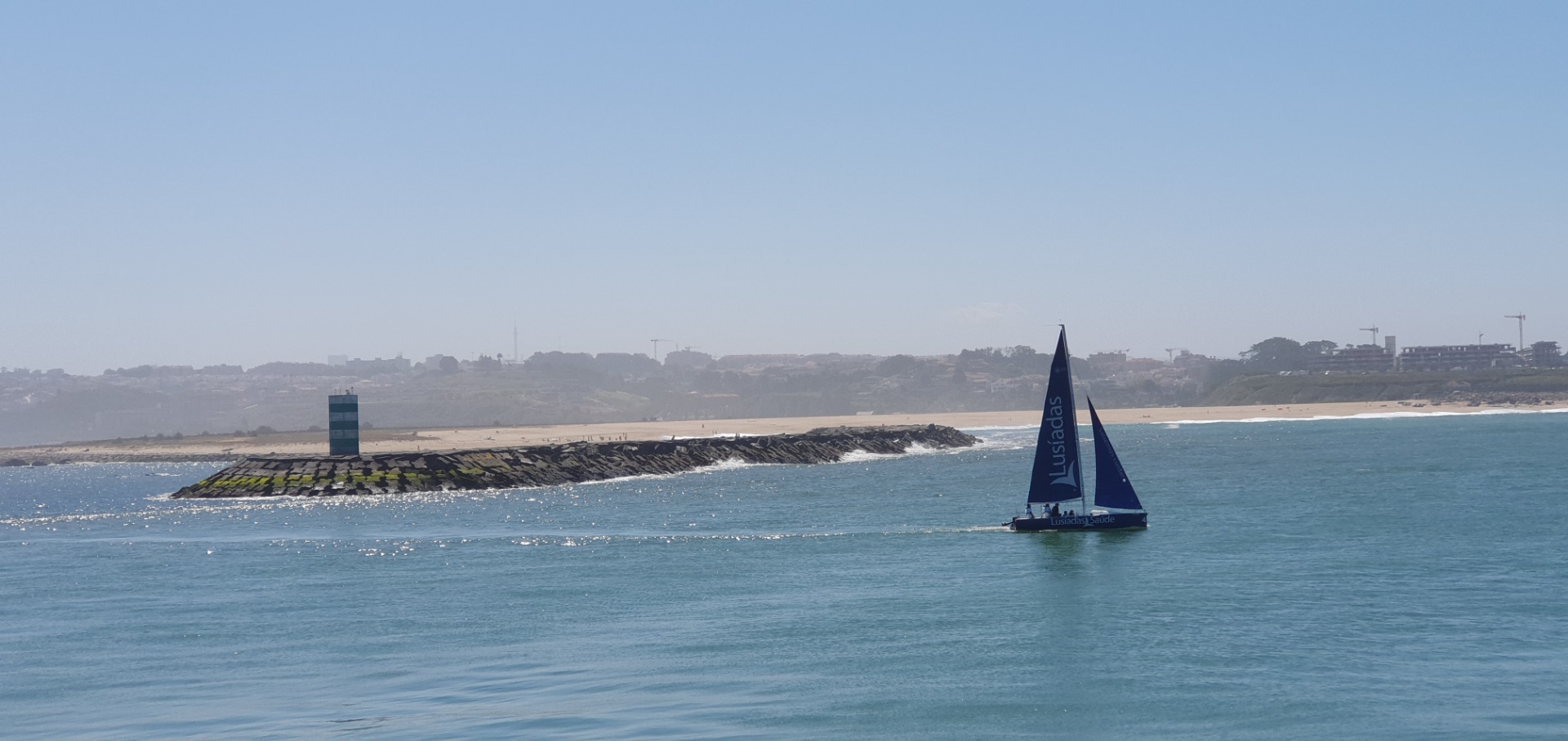 Foz do Douro - Sailboat with “Lusíadas” sails past a breakwater and lighthouse, ocean and sandy coastline in distance.