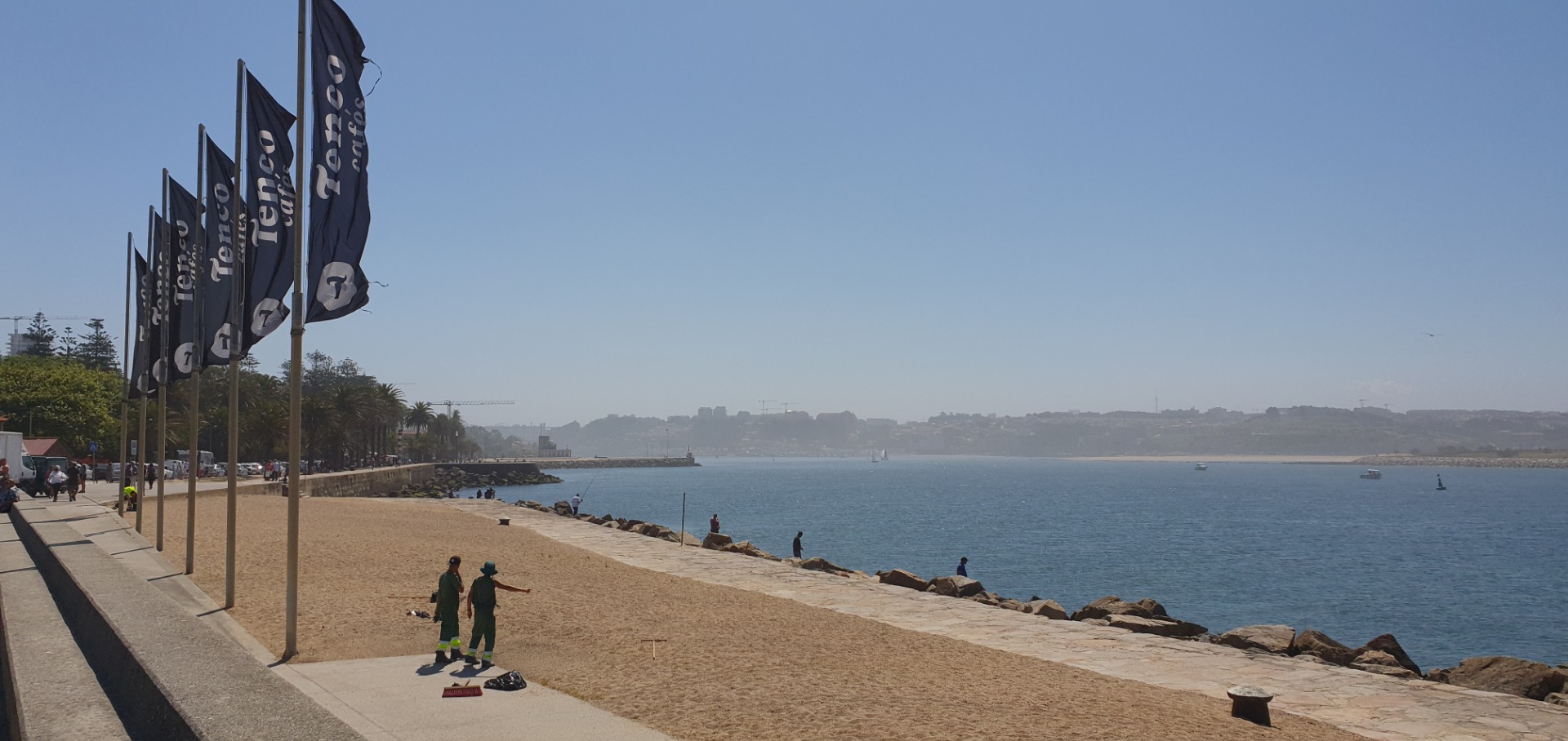 Seaside promenade with dark flags, rocky breakwater, and people walking along calm blue water.