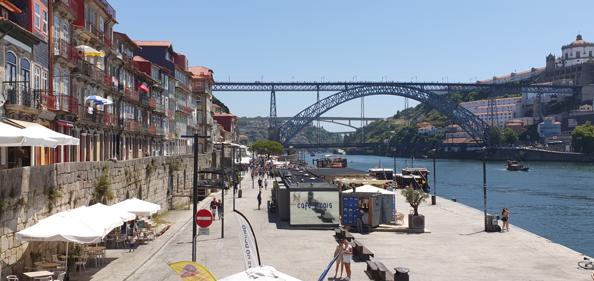 Riverside promenade with cafes and umbrellas, historic buildings, and a large steel arch bridge over calm water.