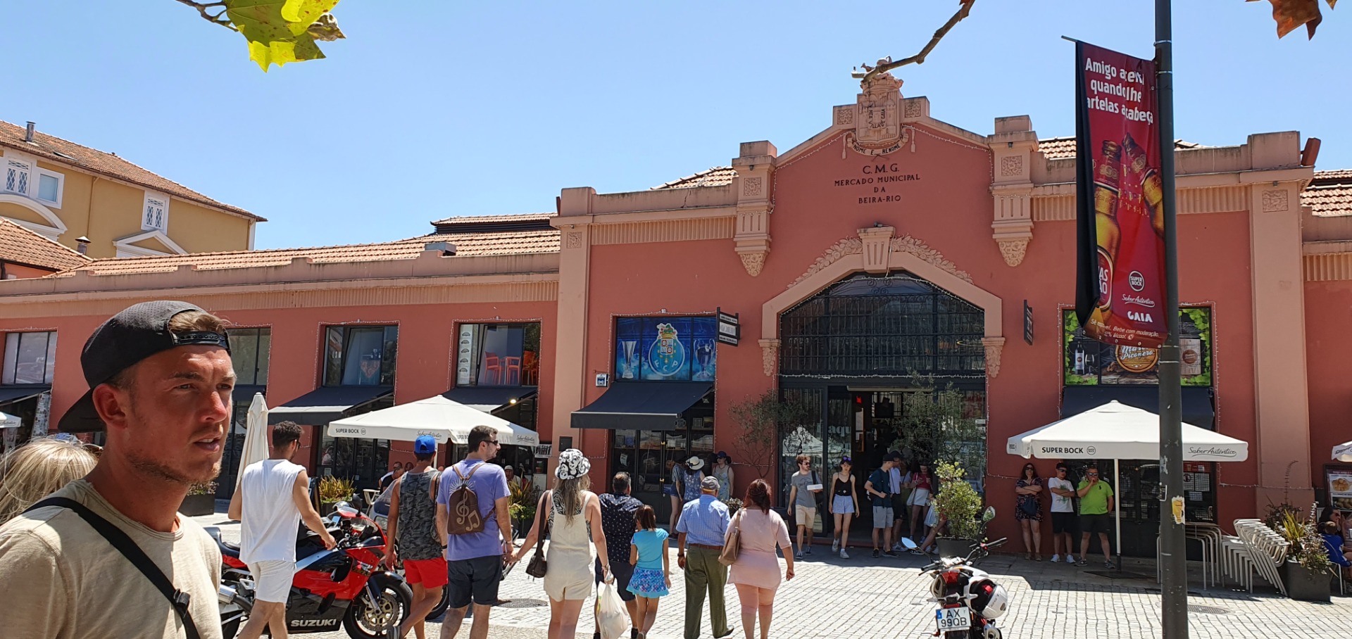 Crowd walks past a pink market building with umbrellas, motorcycles, and a tall street banner.
