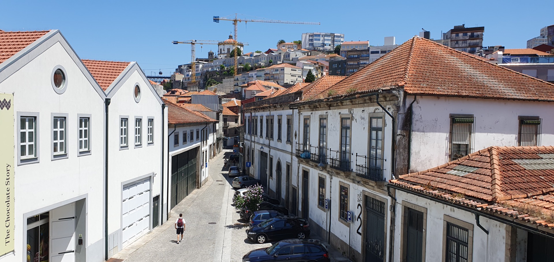 Narrow street between white buildings with red tile roofs, parked cars, and cranes over city skyline.