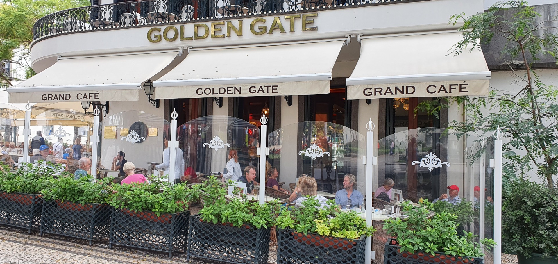 Straßencafé „Golden Gate“ mit weißen Markisen, Gästen im Innenhof und grünen Pflanzen vor dem Eingang.