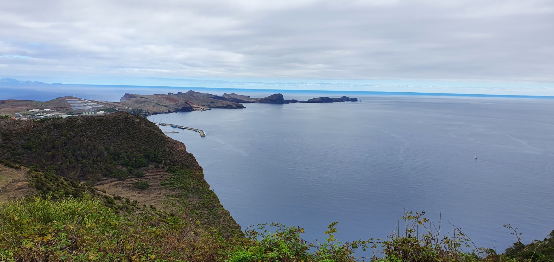 r Ponta de São Lourenço - Weite Küstenlandschaft mit Klippen, Hafenanlagen und ruhigem Meer unter bewölktem Himmel.