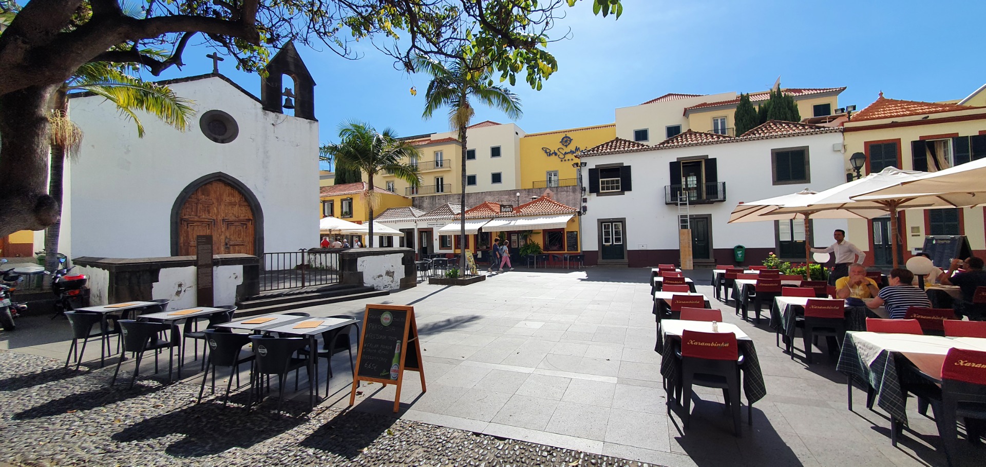 Funchal altstadt - Sonniger Platz mit Café-Tischen, weißer Kapelle mit Glockenturm und bunten Häusern im Hintergrund.