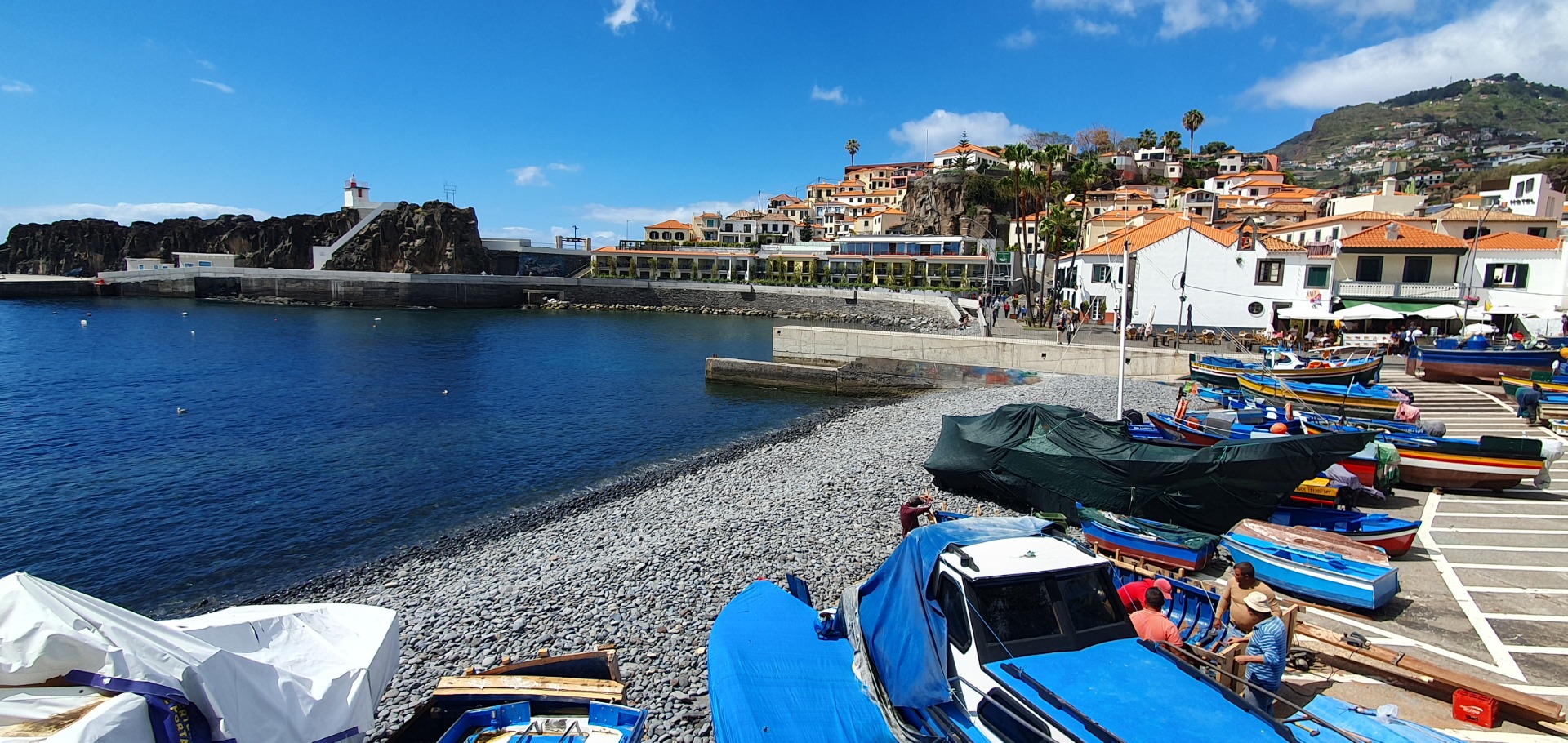 Cámara dos Lobos - Madeira - Harbor waterfront with small boats, rocky shore, white buildings, and orange rooftops under a blue sky.