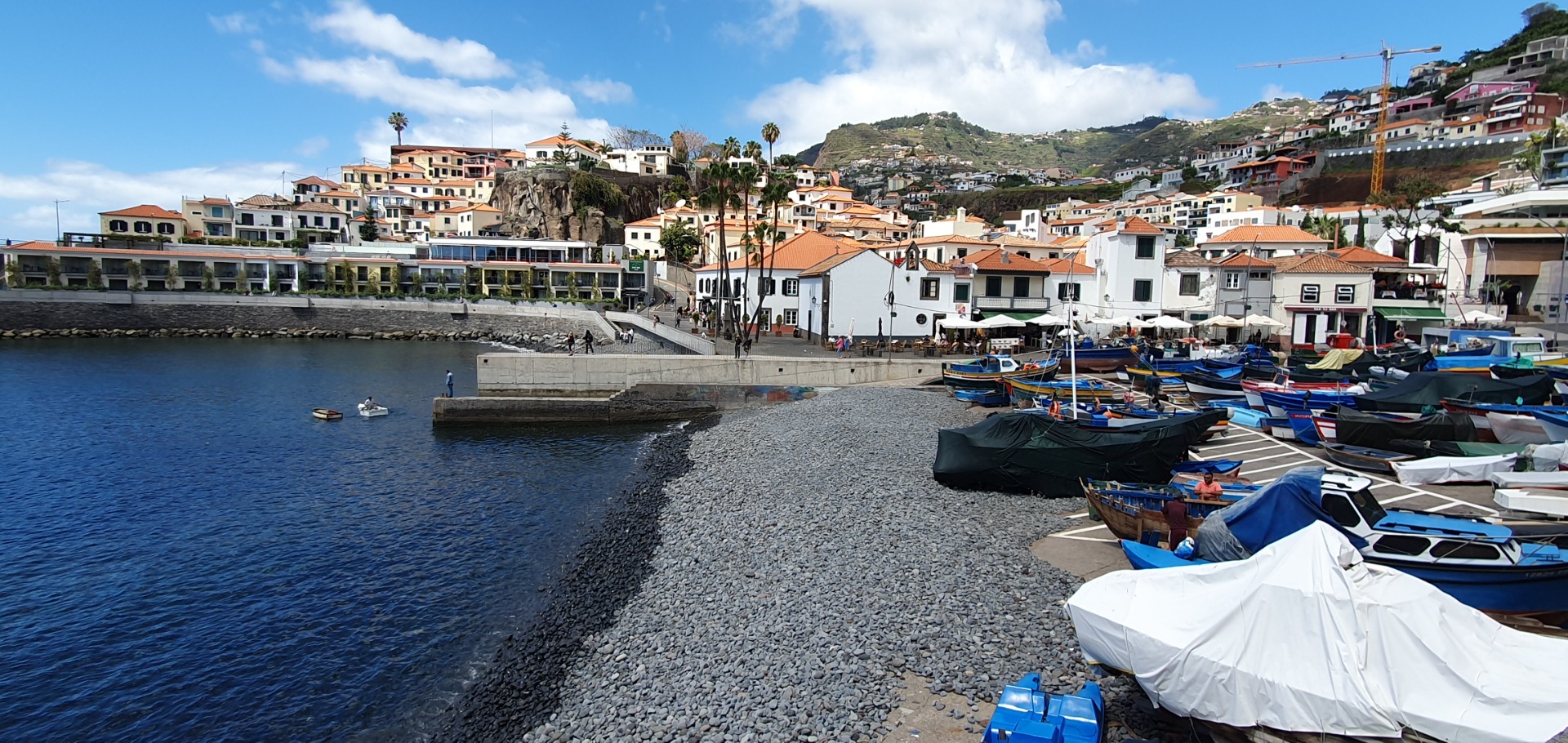 Hafenpromenade Camara dos Lobos mit Fischerbooten, weißen Häusern und bunten Dächern an einer felsigen Küste unter blauem Himmel.
