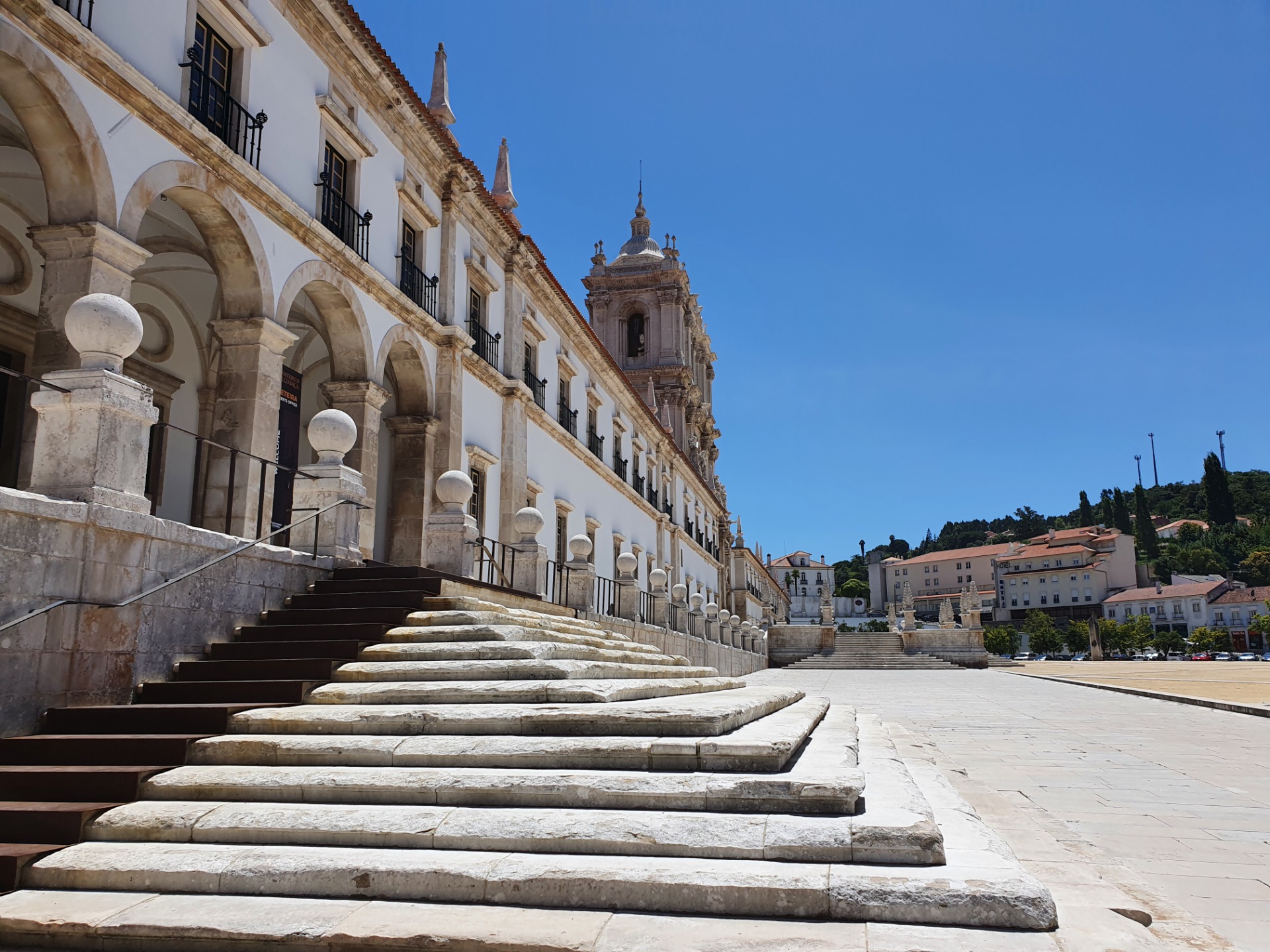 Kloster von Alcobaça - Steinerne Treppen und Arkaden eines historischen Gebäudes, Blick auf eine Kuppelkirche vor blauem Himmel.