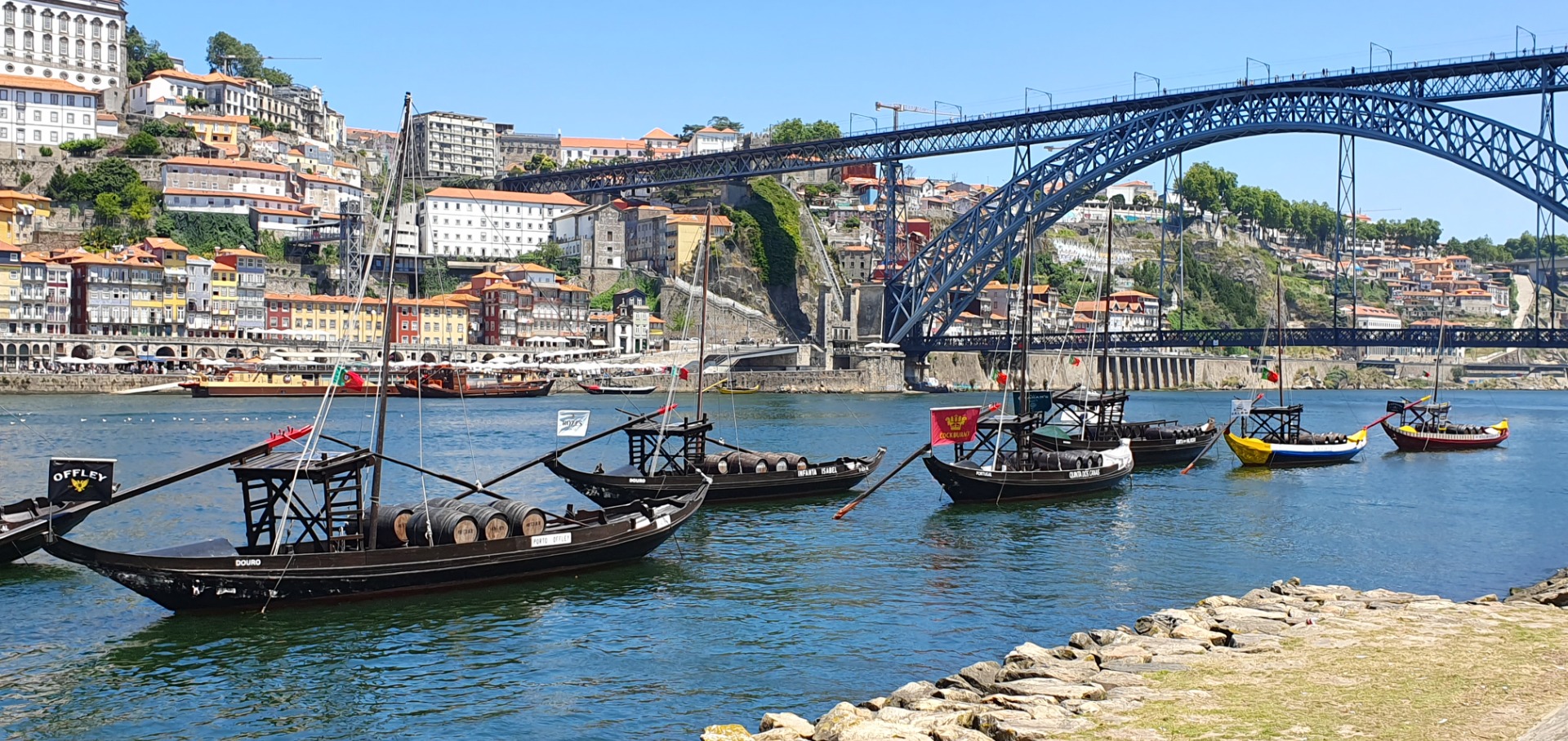 Porto - Douro River Traditional wooden boats moored on a river beneath a steel bridge, with colorful hillside buildings in background.