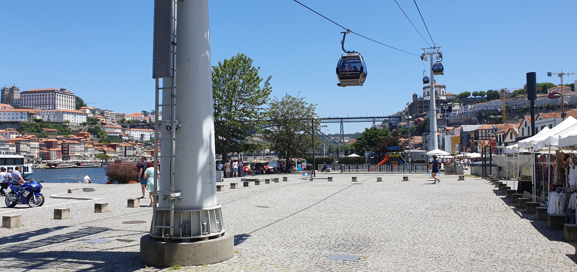 Sunny waterfront plaza with cable car gondolas overhead, people walking, and colorful buildings across the river.