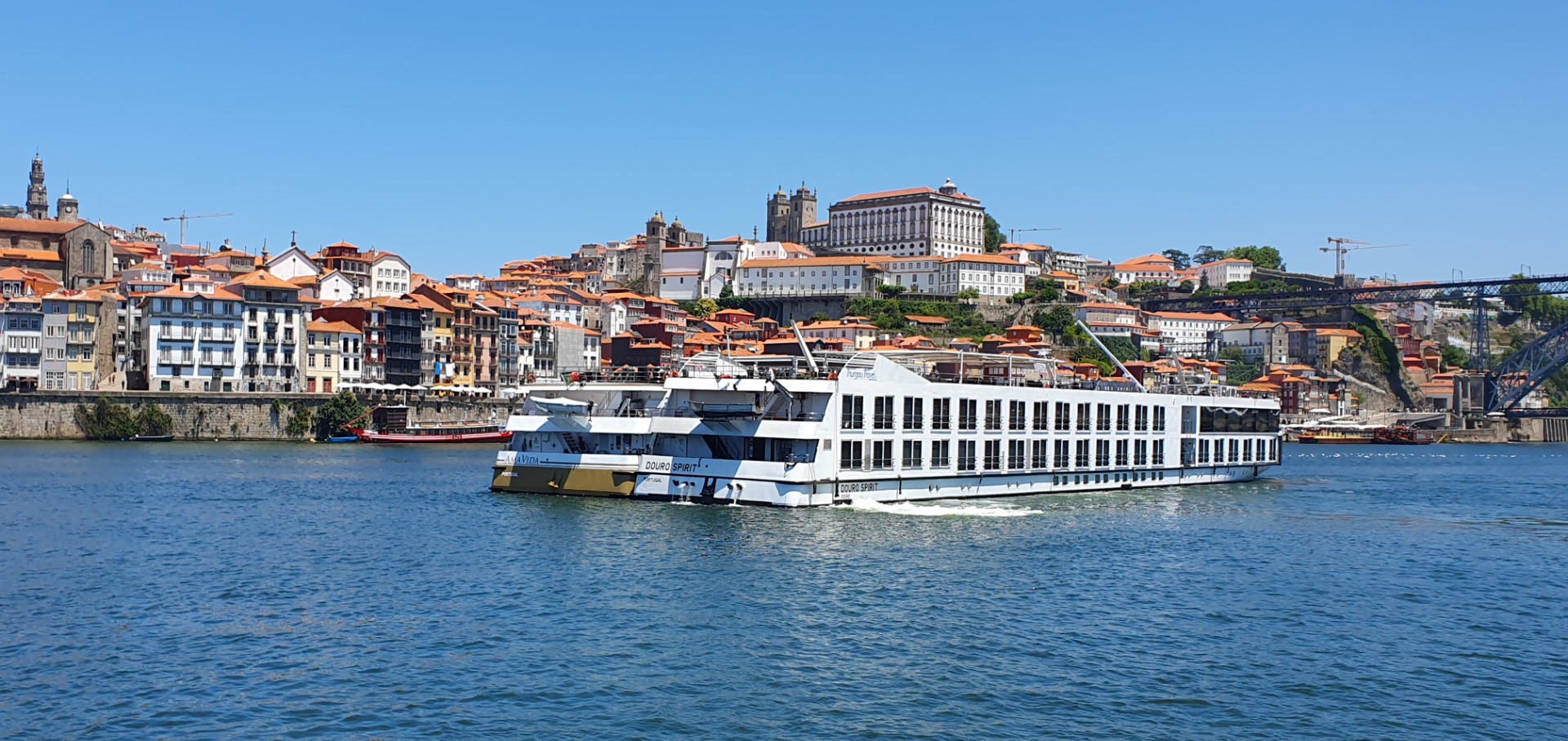 Douro River Cruises- White river cruise ship floats on calm water beside colorful hillside buildings and a blue bridge.